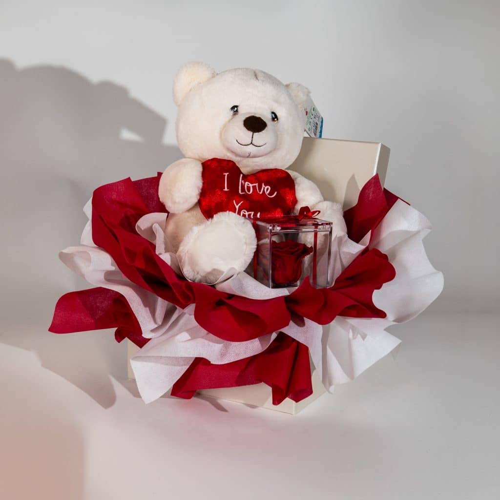 Valentine gift box with teddy bear holding I Love You heart and forever rose in clear box with red and white luxury romantic packaging on neutral background