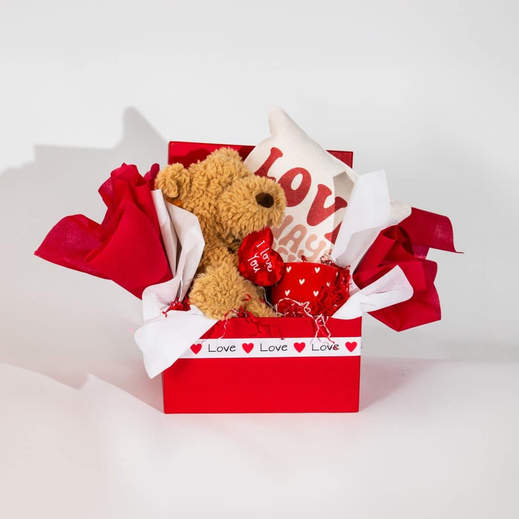 Valentine gift box with teddy bear holding I Love You heart, love cushion and heart mug in red and white romantic packaging on neutral background