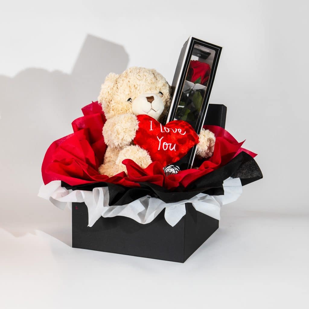 Luxury Valentine gift box with teddy bear holding I Love You heart and forever rose in display case in black red elegant packaging on neutral background