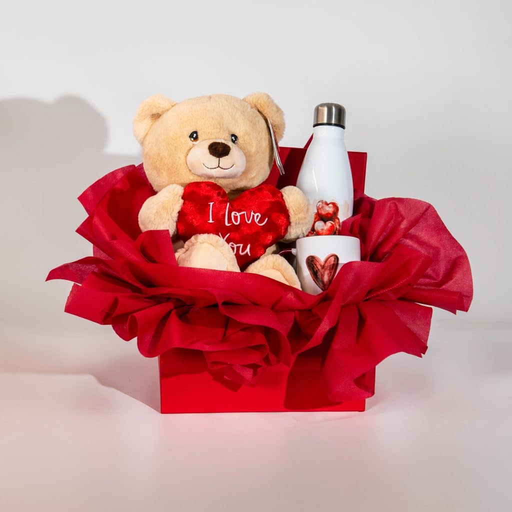 Valentine gift box with teddy bear holding I Love You heart, heart mug and insulated bottle in red luxury romantic packaging on neutral background