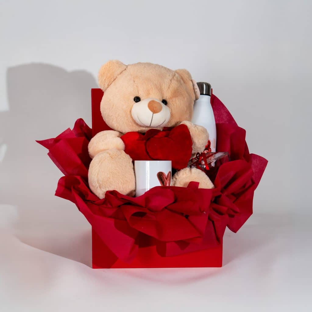 Valentine gift box with teddy bear holding heart, love mug, thermal bottle and chocolates in elegant red romantic packaging on neutral background