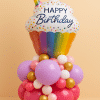 Cupcake-themed birthday balloon arrangement with a large rainbow foil balloon on top and a base of colorful latex balloons in pink, lavender, gold, and white, set against a peach background.
