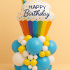 Rainbow cupcake-themed foil balloon arrangement with base of bright blue, yellow, and white latex balloons, creating a fun and vibrant birthday display against a peach background.