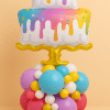 Balloon arrangement featuring a colorful two-tier cake foil balloon with rainbow drips on a golden base, supported by a vibrant base of assorted latex balloons in turquoise, yellow, pink, lilac, and white.