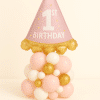 Balloon arrangement featuring a large pink party hat foil balloon with “1st Birthday” text, placed on a base of blush, ivory, and gold latex balloons — perfect for a baby girl’s first birthday celebration.
