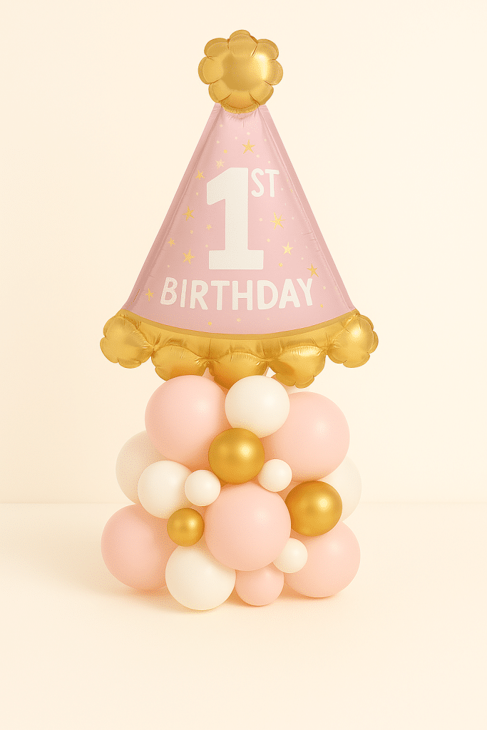Balloon arrangement featuring a large pink party hat foil balloon with “1st Birthday” text, placed on a base of blush, ivory, and gold latex balloons — perfect for a baby girl’s first birthday celebration.