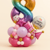 Balloon tower featuring a rainbow number 8 foil balloon with party-themed accents including presents, stars, hat, and "Happy Birthday" message, set on a pink, gold, and lilac base.