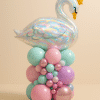 Large iridescent swan foil balloon with gold star crown, sitting atop a deluxe pastel balloon base in blush pink, mint, lavender, and rose gold, decorated with mini balloon garland.