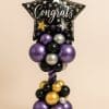 Balloon tower featuring a black star foil balloon with “Congrats” text and multi-color stars, standing on a bold base of metallic purple, gold, black, and silver balloons.