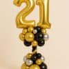 Balloon tower featuring gold foil number 21 balloons, standing on a base of black, gold, and silver balloons with a twisted black-and-gold stem.