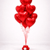 Elegant floor-standing bouquet of red helium foil heart balloons tied with red ribbons on a decorative balloon base, romantic Valentine’s arrangement on a soft neutral background.