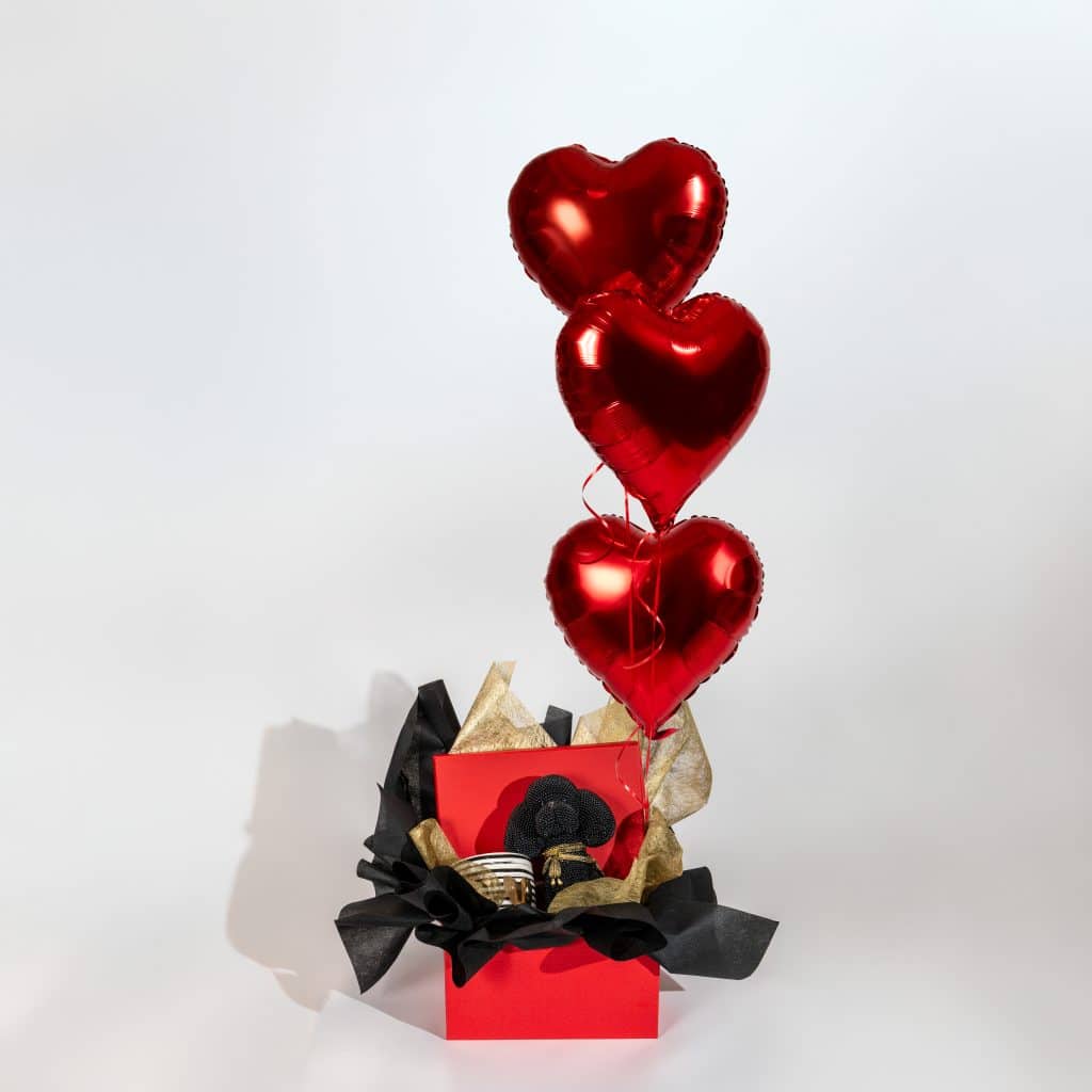 Luxury Valentine gift box with decorative teddy, mug and three red heart foil balloons in red black and gold elegant packaging on neutral background