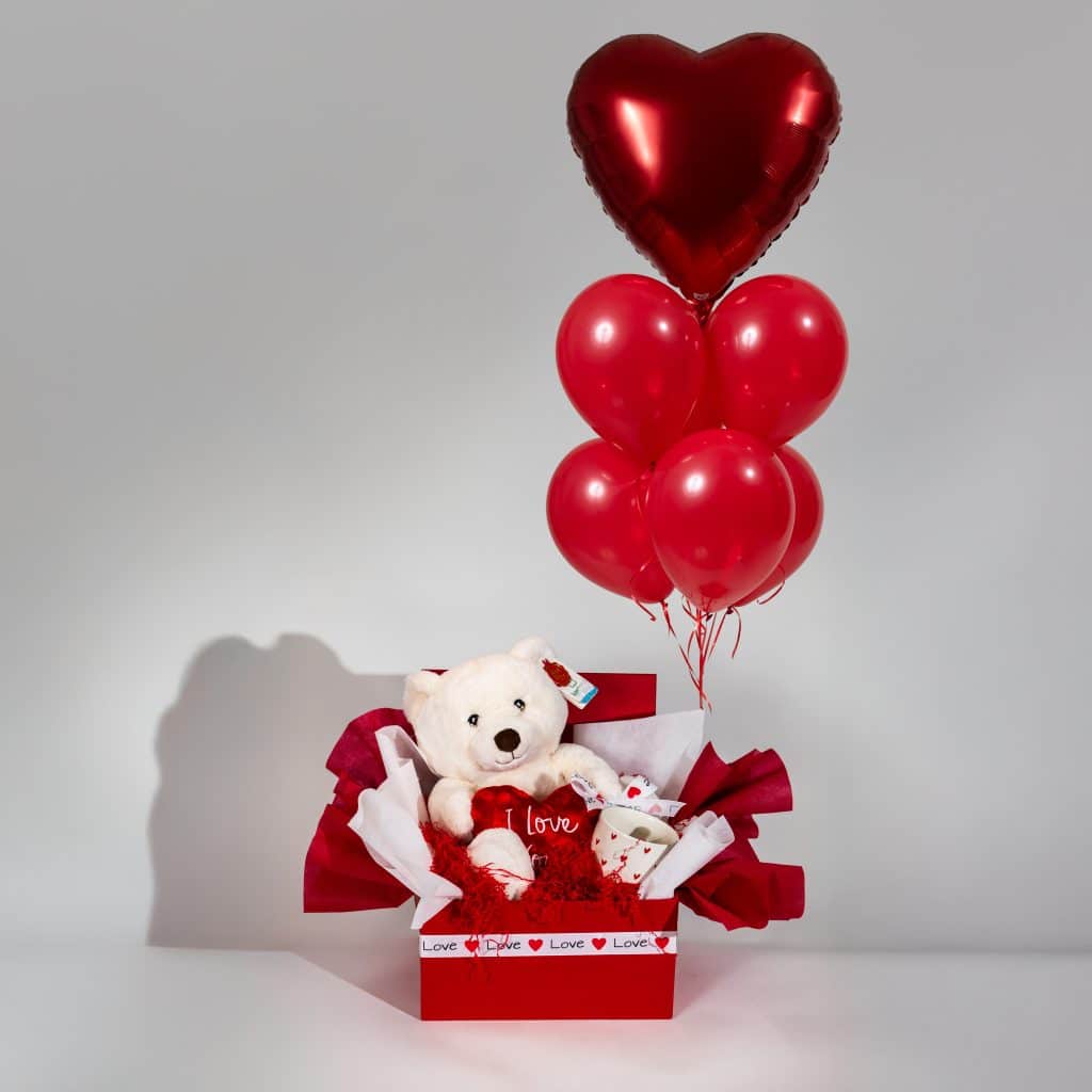Valentine gift box with teddy bear holding I Love You heart, love mug and red balloon bouquet with heart foil balloon in romantic red packaging on neutral background