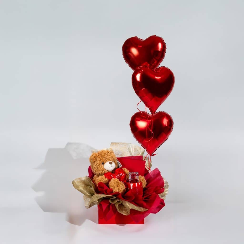 Luxury Valentine gift box with teddy bear holding I Love You heart, red and gold wrapping, chocolate treats and three red heart foil balloons on neutral background