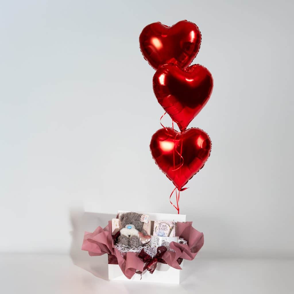 Valentine gift box with teddy bear, sweet treats, blush pink wrapping and three red heart foil balloons on neutral background