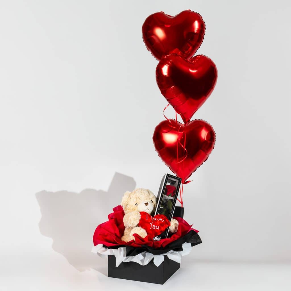 Luxury Valentine gift box with teddy bear holding I Love You heart, preserved forever rose, black and red wrapping and three red heart foil balloons on neutral background