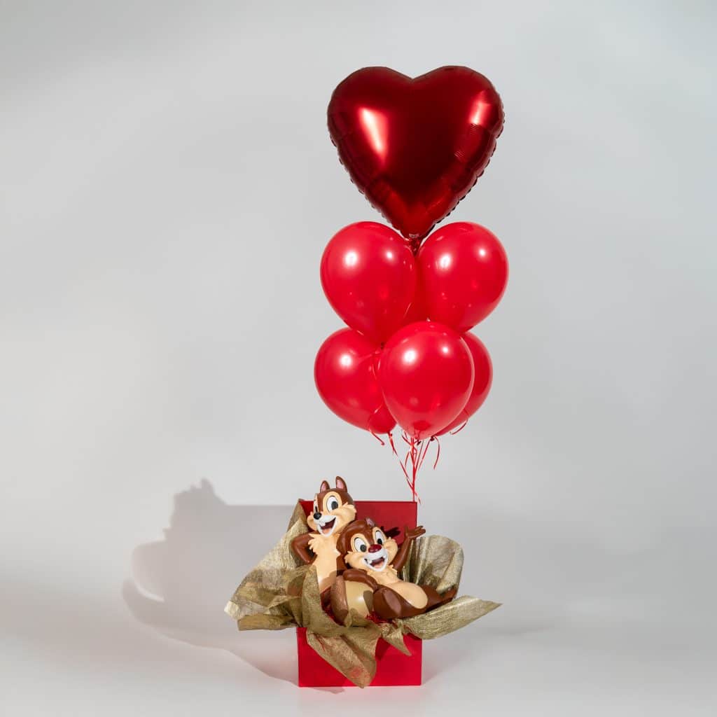 Red heart balloon bouquet rising from a decorative gift box with cute character figures and red helium balloons on a neutral background.