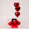 Red gift box with black teddy bear holding a heart and three floating red heart helium balloons on a neutral studio background.