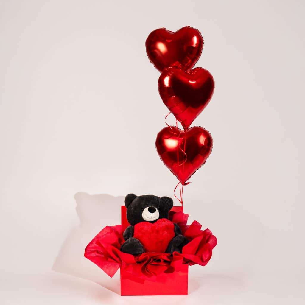 Red gift box with black teddy bear holding a heart and three floating red heart helium balloons on a neutral studio background.