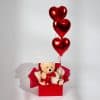 Valentine’s gift box with teddy bear holding heart cushion, ceramic mug, and three red heart foil balloons on a neutral background.